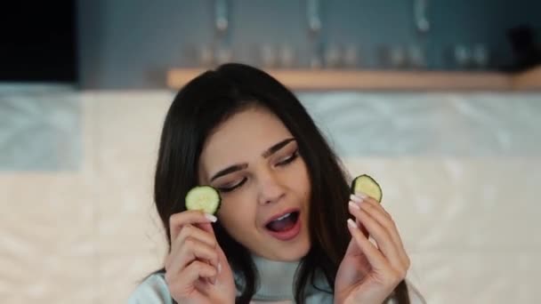 Mujer joven tiene muecas y hacer caras con rodajas de pepino mientras se cocina en la superficie de la cocina. Hay pepinos, tomates, pimientos, huevos, hojas de lechuga a su lado. — Vídeos de Stock