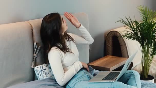 Des technologies qui aident les gens à communiquer. Jeune femme brune est assise sur le canapé et utilise un smartphone et un ordinateur portable. Le modèle féminin a une conversation au téléphone tout en surfant sur Internet. — Video