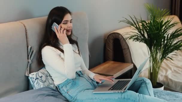 コミュニケーションを支援する技術。若いブルネットの女性はソファに座ってスマートフォンやラップトップを使用しています。女性モデルはインターネットをサーフィンしながら電話で会話をしています. — ストック動画