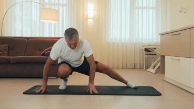 Yavaş çekim. Yaşlı adam yan atlama egzersizi yapıyor. Formda vücut ısısı olan son sınıf erkek atlet, evde bacak esnetme egzersizi yapıyor. Kendine iyi bak. Arka planda modern ışık odası..