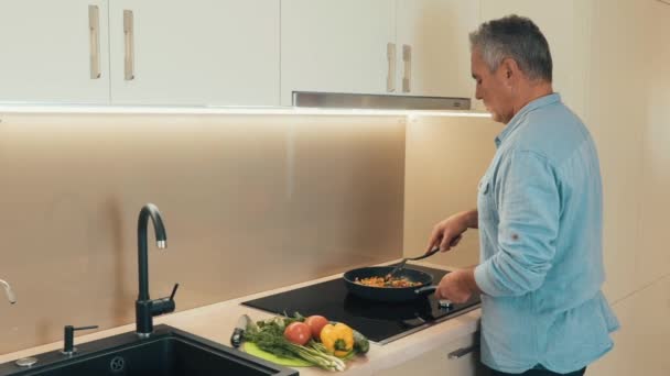 Homem concentrado em roupas casuais fica junto ao fogão e ensopa legumes em uma frigideira. Homem maduro preparando comida saudável para o jantar para sua família. Conceito de alimentação saudável. filmagens em câmera lenta — Vídeo de Stock