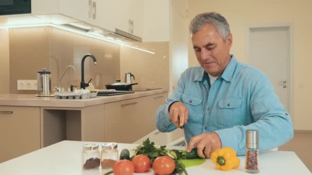 Voksen mand hugger grøntsager og greens på et køkkenbord. Klip udsigten. Agurker, tomater, peberfrugter, courgetter og krydderikrukker på rent hvidt bord. Madlavning med glæde koncept. Optagelser med langsom bevægelse – Stock-video