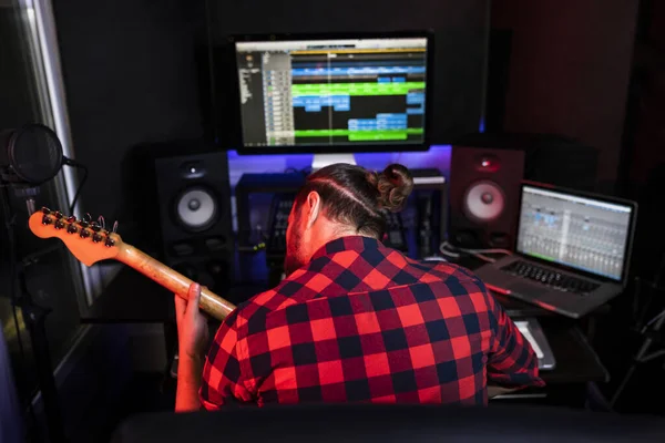 Hipster άνθρωπος παίζει κιθάρα και sining νέο τραγούδι του σε στερεοφωνικό στούντιο για να ηχογραφήσετε το ολοκαίνουργιο κομμάτι. Χρησιμοποιώντας μίξερ, στερεοφωνικό σύστημα, και πρόγραμμα παραγωγής — Φωτογραφία Αρχείου