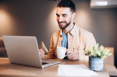 Memleketten çalışan neşeli, serbest çalışan bir adam. Çekici genç adam gülümsüyor, modern laptopunu kullanıyor ve video aracılığıyla konuşuyor. Arka planda rahat bir mutfak. Kâğıtlar ve kahve kupası masanın üstünde..