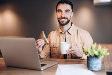 Esmer, serbest çalışan bir adam kahvaltı yapıyor ve kameraya bakıyor. Biraz daha kafein. Günlük giysiler içinde dizüstü bilgisayar ve programlama kullanan bir adam. Uzaktan çalışma konsepti. Mutlu çekici adam kameraya bakıyor..