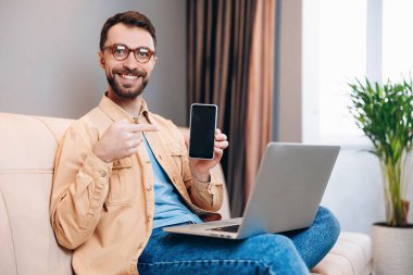 Erkek serbest çalışan, evden çalışmaktan mutlu. Parlak gülüşlü, akıllı bir adam bir elinde akıllı telefonu tutuyor ve diğerini işaret ediyor. Dizüstü bilgisayarında. İçi aydınlık oturma odası..