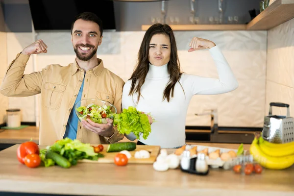 Cute vegetarian couple is showing how healthy and strong they are. Happy vegan people are cooking salad using only nutrition and healthy food