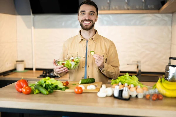 Handsome guy with beard and nice smile is cooking salad. He probably likes healthy lifestyle and eating fresh food. Young vegetarian man is cooking in kitchen indoor