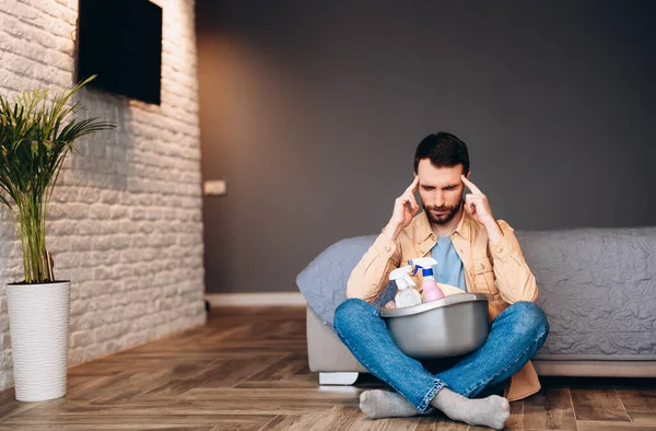 Hombre estresado mirando decepcionado antes de limpieza de la casa, sentado en el suelo con suministros de limpieza, espacio de copia. — Foto de Stock