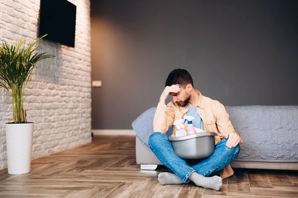 Hombre estresado mirando decepcionado antes de limpieza de la casa, sentado en el suelo con suministros de limpieza, espacio de copia — Foto de Stock