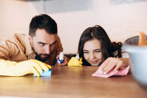 Happy couple doing house cleaning in modern kitchen at home. Close up of young man and woman washing counter top with detergent and cloth with wife.