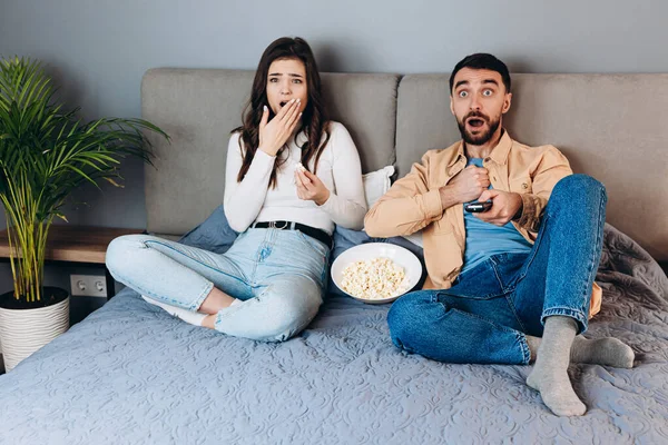 Omg何ですか?。甘いカップルは、屋内の家の中でポップコーンボックスに座ってソファで終わる予期せぬスリラー映画を驚かせた隔離された自由な時間の時計映画を持っています。素敵なショックを受けたカップル見ますテレビ — ストック写真