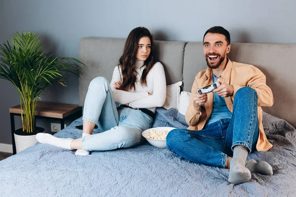 甘いカップルは隔離された自由な時間がビデオゲームをする。彼女は好きではないと失望感退屈しながら彼あります楽しいですとともにポップコーンボックス座ってソファで家の中に — ストック写真