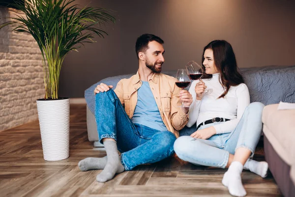 Νεαρό ζευγάρι κάθεται στο πάτωμα με κόκκινο κρασί στην κρεβατοκάμαρα και γιορτάζει το νέο τους διαμέρισμα. — Φωτογραφία Αρχείου