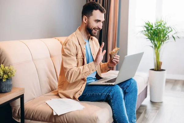 Man speaking by videocall, eating sandwich and sitting on sofa in his cozy living room. Man communicating with friends using his laptop and smiling. Remote worker.