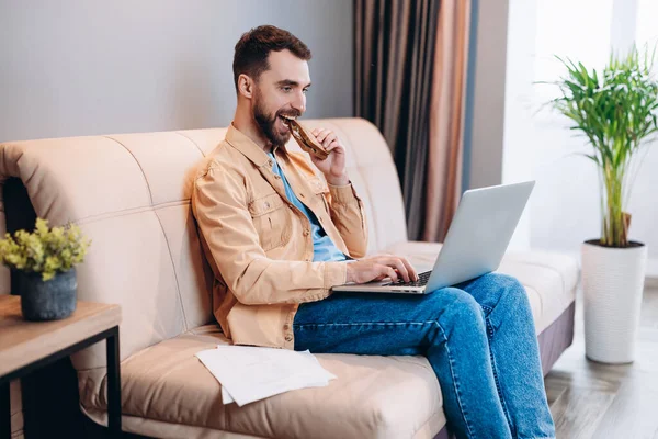 Man watching funny videos on laptop. Good looking young man in casual clothes eats lunch while working from home. Quarantine concept. Living room on background.