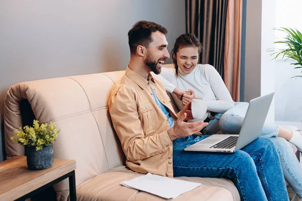 若いカップルは一緒に面白いビデオに見えます.カジュアルな服を着た男と女は、現代のリビングルームの革のソファに座って、人間のラップトップで何かを笑う。家で幸せな家族の時間. — ストック写真