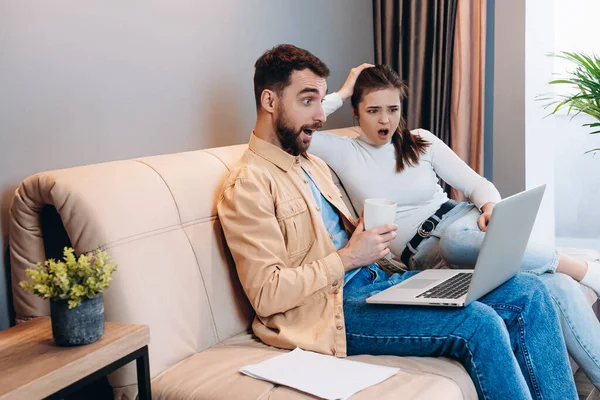 見てみろよ。オレンジ色のシャツと青いジーンズの良い外見の男と白いタートルネックの魅力的な若い女性が一緒にノートパソコンの画面上で衝撃的な何かを見た。スマートリビング室内. — ストック写真