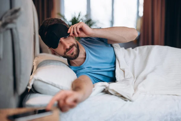 Joven con barba despertando en la cama se acerca para apagar la alarma en el teléfono móvil. — Foto de Stock