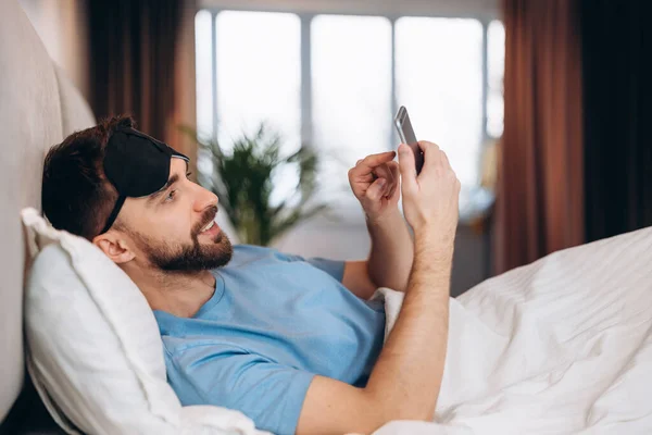 Joven hombre atractivo acostado en la cama revisando sus mensajes en su teléfono en el día libre perezoso de la mañana. — Foto de Stock