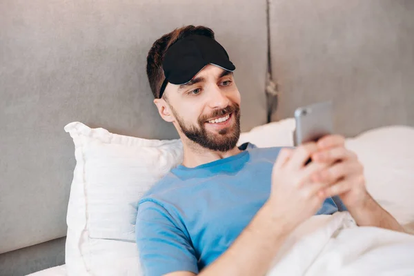 Joven hombre atractivo sonriendo y acostado en la cama revisando sus mensajes en su teléfono en la mañana perezoso día libre. — Foto de Stock
