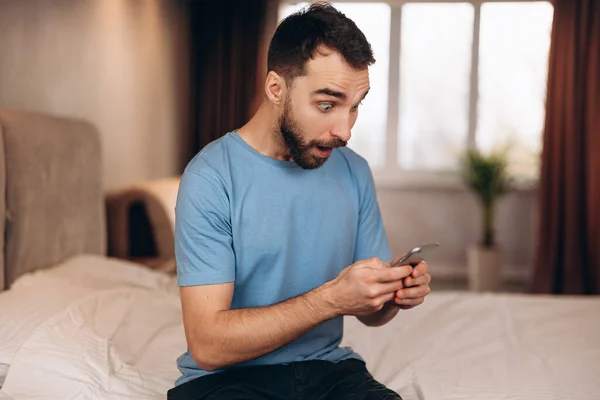 Joven con barba sorprendido mientras recibía el mensaje por teléfono mientras estaba sentado en la cama. — Foto de Stock