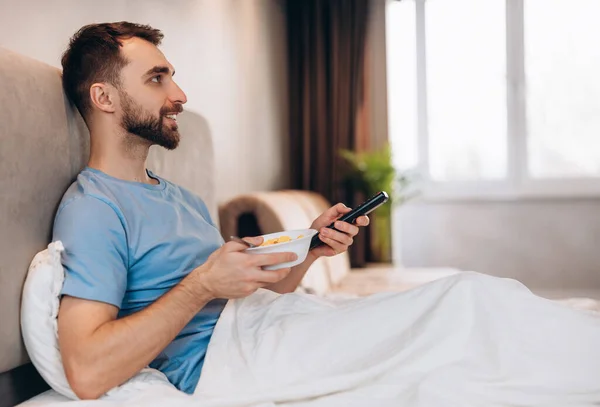 Jóképű fiatalember szakállal az ágyban reggelizik és tévét néz. hétvégi lusta szabadnap. — Stock Fotó