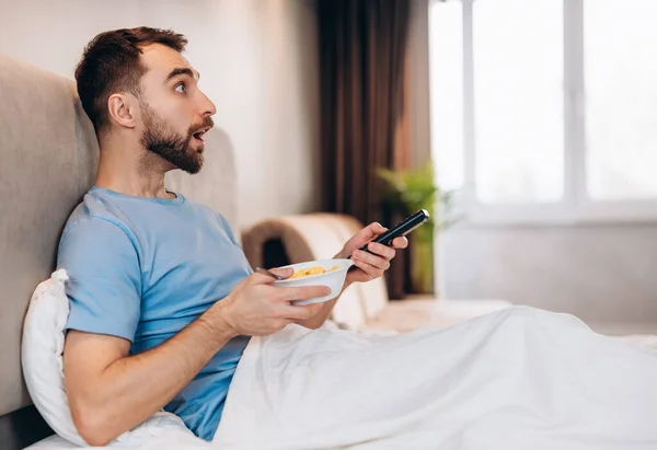Vonzó fiatalember szakállal reggelizik az ágyban, és meglepetten néz, miközben híreket néz.. — Stock Fotó