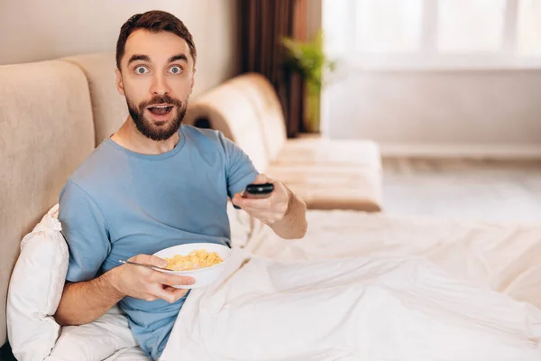 Vonzó fiatalember szakállal reggelizik az ágyban, és meglepetten néz, miközben híreket néz. — Stock Fotó