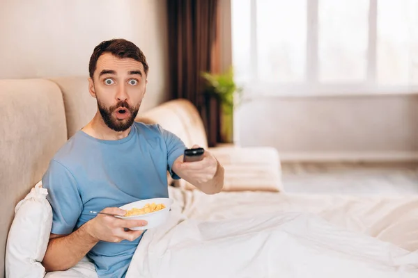 Vonzó fiatalember szakállal reggelizik az ágyban, és csalódottan néz, miközben híreket néz.. — Stock Fotó