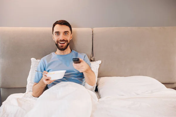 Vonzó fiatalember szakállas eszik reggeli az ágyban, és úgy néz ki, boldog tévéműsorok nézése közben. — Stock Fotó
