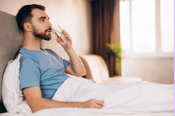 Guapo joven con barba sonriendo agua potable antes del desayuno en la cama mira a la cámara. fin de semana día libre perezoso. — Foto de Stock