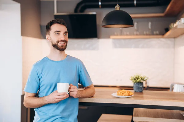 Daily life of young man in kitchen during breakfast holding a cup of coffee. Attractive male working from home, telecommuting using remote internet job online communication on