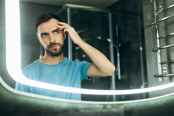 Молодой человек с бородой в синей футболке ищет прыщ на лице в ванной зеркало акне проблема. — стоковое фото