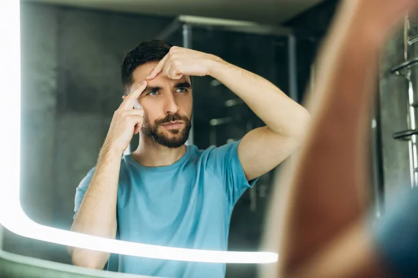 Fiatal férfi szakállal kék pólóban szorító pattanás fürdőszobában tükör akne probléma. — Stock Fotó