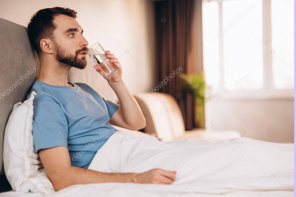 Handsome young man with beard smiling drinking water before breakfast in bed look at camera. weekend lazy day off.