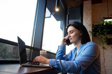 Uzaktan kumanda sırasında telefonla konuşan serbest çalışan bir kadın. Sıcak kahve dükkanı atmosferi. Genç bayan projenin detaylarını müşteriyle tartışıyor. Arka planda kafe ya da restoran.