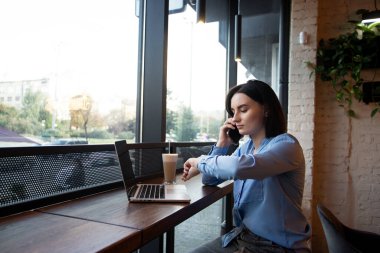 Serbest çalışan kadın saatine bakıyor ve müşteriyle bir şeyler konuşuyor. Uzaktan çalışma konsepti. Arka planda kafe ya da restoran var. Küçük bir zaman kavramı. Bir fincan lezzetli latte..