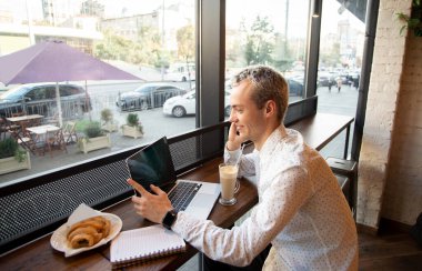 Genç serbest çalışan bir telefon görüşmesinden memnun. Uzaktan çalışma konsepti. Arka planda kahve evi var. Lezzetli kruvasan ve bir fincan latte. Bir kafede çalışan serbest çalışan erkek programcı..