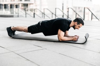Plank it Büyük şehir konsepti eğitimi. Siyah spor giyimli genç adam asfalt yollarda egzersiz yaparken kalas pozisyonu alıyor. Sıkı çalışma. Bencil bakım kavramı.