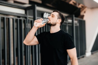 Kapalı gözlerle koştuktan sonra plastik şişeden su içen adam. Sıkı çalışma. Kendine dikkat et konsept. Biraz daha su iç. Büyük şehir konseptinde spor.