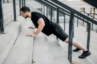 İyi yapılı genç bir adam merdivenlerden yukarı itiyor. Yan görüş. Siyah saçlı, siyah spor giyimli, spor ayakkabılı, sokakta spor yapan bir atletik. Formda bir vücuda sahip olmak için idman yapan konsantre bir adam..