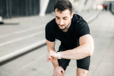 Adam koşucu dinleniyor ve saatine bakıyor. Kardiyo yük kontrol konsepti. Çekici esmer adam sabahları siyah spor kıyafetleriyle koşuyor. Arka planda gri şehir.
