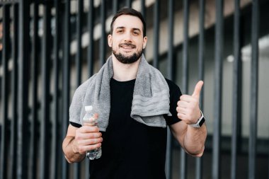 Akıllı telefonları olan neşeli bir adam başparmak kaldırıyor. Biraz daha su iç. Şehir sporu konsepti. Sabah antrenmanından sonra omuzlarında havlu olan bir adam dinleniyor. Jogging konsepti.