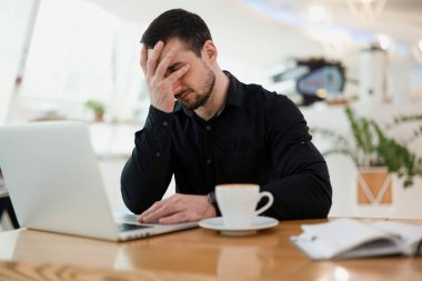 Serbest çalışan adam facepaling. Kahvehaneden uzak bir iş. Siyah gömlekli genç işadamı modern laptopunun önünde facepalm hareketi yapıyor. Backgorund 'daki kafede..