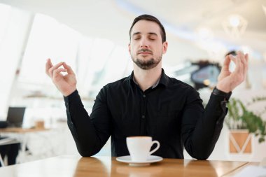 Sakin ol ve bir fincan espresso iç. Adam meditasyon yapıyor çünkü çok fazla kafein aldığı için panik atak geçirmiş. Arka planda kahve dükkanı var. Şık adam..