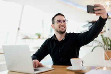 Gülümseyen adam konsantrasyon problemleri üzerinde çalışması gerekirken selfie çekiyor. Adam bugün çalıştığı yeri arkadaşlarına gösteriyor. Sosyal medya saçmalıkları. Arka planda açık kahve evi.