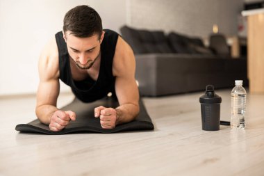 Yoga minderiyle egzersiz yaparken konsantrasyonlu adam kalas pozisyonu alıyor. Su şişeleri yakınlarda duruyor. Arka planda modern bir iç mekan. Siyah giysili adam evde spor yapıyor..