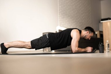 Plank it Evde eğitim. Kendine güvenen genç adam siyah spor giysiler giyiyor ve tavan arasında yerde egzersiz yaparken kalas pozisyonu alıyor. Sıkı çalışma..