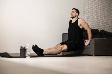 Çekici adam kanepeden itiyor. Siyah spor giyim. Kollarını güçlendirmek için sıkı çalışan bir adam. Plastik su şişesi. Siyah yoga minderi. Neşeli erkek sporcu.